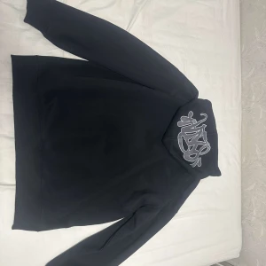 Svart hoodie med graffiti-tryck - Svart hoodie med coolt vitt graffiti-tryck på huvan. Tröjan har huva med snörning och en klassisk känguruficka framtill. Perfekt för dig som gillar streetwear och vill ha en stilren men ändå unik look.