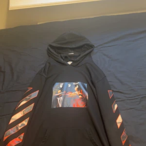 Svart Off-White hoodie med tryck - Svart hoodie från Off-White med stor grafisk print på ryggen och färgstarkt motiv på bröstet. Ärmarna har röda och vita diagonala ränder. Klassisk huva och känguruficka framtill. Perfekt för dig som gillar streetwear och vill sticka ut.