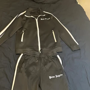 Svart zip hoodie från Palm Angels - Svart hoodie från Palm Angels med vita detaljer och dragkedja framtill. Tröjan har vita ränder längs ärmarna och två fickor med dragkedja. Snygg streetwear-stil med logga på bröstet. Perfekt för dig som gillar sportig och trendig look.