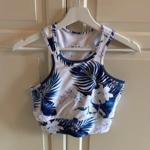 Blommig träningstopp från Stronger - Säljer en snygg sporttopp från Stronger i vitt med blått och rosa blommönster. Toppen har brottarrygg och bred resår nedtill för extra stöd. Materialet är mjukt och stretchigt syntet som andas. I fint skick, förutom att trycket på nacken är slitet (se bild). Storlek M. 