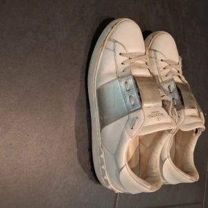 Valentino Open sneakers i vitt och silver - Snygga Valentino Open sneakers i vitt skinn med bred silverfärgad detalj över vristen. Klassisk rund tå, snörning och platt sula. Märkeslogga på plösen och coola nitar baktill. Perfekta för dig som vill sticka ut med stilrena men ändå unika sneakers. Lite skador på höger fram sidan om man kollar nära 
