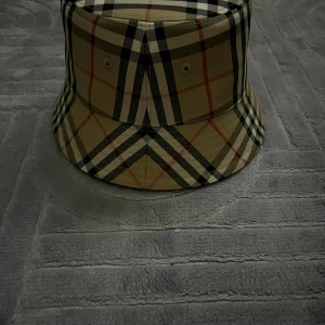 Burberry bucket hat i klassiskt rutigt - Snygg bucket hat från Burberry i deras ikoniska beigea rutmönster med svarta, vita och röda detaljer. Hatten är tillverkad i bomull och har en avslappnad form som ger en cool vibe till din outfit. Perfekt accessoar för att sticka ut i mängden.
