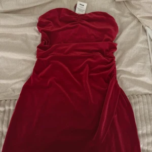 Röd sammetsklänning DK Harper - Röd axelbandslös sammetsklänning från DK Harper i kort modell. Klänningen har en snygg drapering i midjan och en figurnära passform. Perfekt för dig som vill sticka ut med en elegant och trendig look.