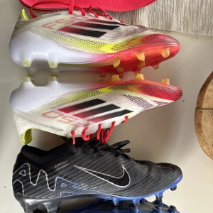 Adidas och Nike fotbollsskor - Två par fotbollsskor: ett par Adidas i vitt med svarta och röda detaljer samt gula och röda inslag, och ett par Nike i svart med vita och blå detaljer. Båda har snörning och dobbar för spel på gräs. Perfekta för dig som vill sticka ut på planen.