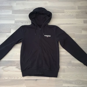 Hoodie - Napapijri - Svart hoodie från Napapijri med dragkedja och huva. Snyggt tryck med logga och norsk flagga på bröstet. Mjuk insida och två fickor framtill. Perfekt för chill dagar och streetstyle. Klassisk passform och långärmad.