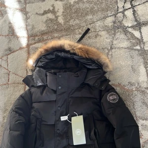 Svart dunjacka från Canada Goose - Svart dunjacka från Canada Goose med stor huva och snygg pälsdetalj. Jackan har broderad logga på ärmen, flera fickor och stängs med både dragkedja och knappar. Materialet är polyester och dun, perfekt för kalla vinterdagar.
