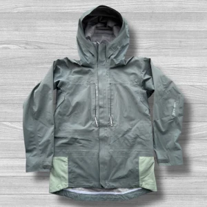 Houdini regnjacka  - Snygg grå regnjacka från Houdini i dammodell med huva och dragkedja framtill. Jackan har snörning vid halsen, praktiska fickor och ljusgröna detaljer vid nederkanten. Perfekt för blöta dagar och outdoor-äventyr.