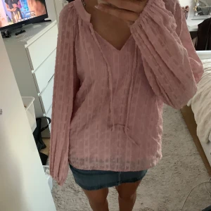 Ljusrosa blus med knyt och v-ringning - Säljer en ljusrosa blus med v-ringning och knytband framtill. Blusen har långa, luftiga ärmar med resår vid handlederna och ett diskret rutigt strukturmönster i tyget. Perfekt för dig som gillar en romantisk och avslappnad stil.