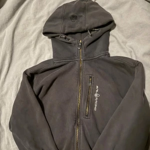 Svart hoodie från Sail Racing - Svart hoodie från Sail Racing med dragkedja framtill och en extra bröstficka med dragkedja. Tröjan har huva med snörning och diskret logga på bröstet. Perfekt för dig som gillar en sportig och stilren look. Lite solblekt men nästan ny, köpt för 1500kr