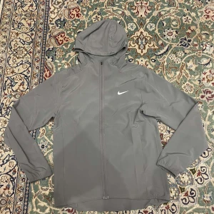 Grå Nike vindjacka med huva - Grå vindjacka från Nike med huva och hel dragkedja framtill. Jackan är tillverkad i 100% polyester och har Dri-FIT-teknologi för att hålla dig torr. Elastiska muddar vid ärmslut och diskret Nike-logga på bröstet. Perfekt för aktiva dagar.