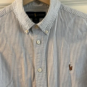 Randig skjorta från Ralph Lauren - Snygg skjorta från Ralph Lauren i vitt och blått med tunna vertikala ränder. Klassisk button-down krage, broderad logga på bröstet och långa ärmar. Skjortan är i bomull och har en clean, tidlös look med knappar framtill.