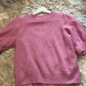 Rosa stickad tröja från Vero Moda - Superfin rosa stickad tröja från Vero Moda med korta puffärmar och ribbade muddar. Tröjan har en rund halsringning och är gjord i ett mjukt material som känns skönt mot huden. Perfekt för dig som gillar en söt och trendig look.