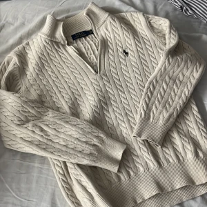 Beige kabelstickad tröja Polo Ralph Lauren - Kabelstickad Beige tröja från Polo Ralph Lauren med halv dragkedja och broderad logga på bröstet. Tröjan har ribbade muddar, hög krage och är långärmad. Perfekt för lager-på-lager och snygg till jeans eller kjol.