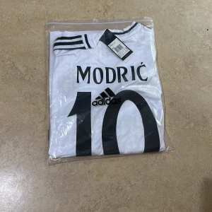 Real Madrid Modric 10 Adidas fotbollströja - Säljer en helt ny officiell Real Madrid 25/26 fotbollströja med Modric och nummer 10 på ryggen. Tröjan är vit med svarta detaljer, klubbmärke på bröstet, Champions League-märke på ärmen och sponsortryck framtill. Tillverkad i lätt och ventilerande material.