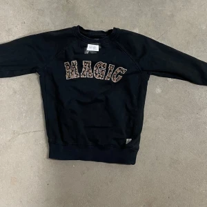 Svart sweatshirt MAGIC från 157 - Svart sweatshirt från 157 med coolt MAGIC-tryck i leopardmönster på bröstet. Tröjan har rund hals och långa ärmar, perfekt för dig som gillar streetstyle och vill sticka ut lite extra. Skön och mjuk bomullskänsla.