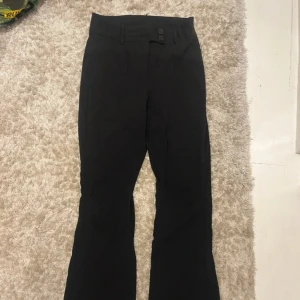 Svarta bootcut kostymbyxor från ONLY - Svarta kostymbyxor från ONLY med bootcut-snitt och hög midja. Byxorna har två knappar i midjan, bälteshällor och fickor med lock bak. Snygg och stilren modell i mjukt material som passar perfekt till en trendig look. Använd mycket få gånger! 