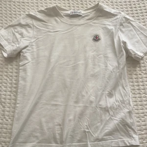 Vit t-shirt från Moncler - Snygg vit t-shirt från Moncler med klassisk rund hals och korta ärmar. T-shirten har en liten broderad Moncler-logga på bröstet och är gjord i mjuk bomull. Perfekt för dig som gillar stilrena och exklusiva plagg.