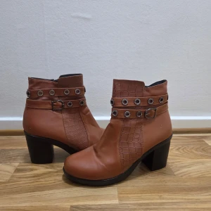 Bruna boots med krokopräglad detalj - Snygga bruna boots med krokopräglade paneler och två remmar med silvriga nitar och spänne runt ankeln. Skorna har en stadig klack och dragkedja på insidan för enkel påtagning. Perfekta för dig som vill sticka ut lite extra.