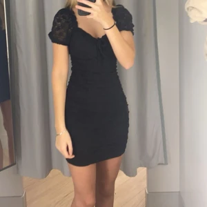 Svart kort klänning från Divided XS - Supersöt svart kort klänning från Divided i storlek XS. Klänningen har puffärmar, knut fram och är sydd i ett lätt strukturerat tyg med diskret mönster. Snygg rynkning längs sidorna och en feminin passform som framhäver siluetten.