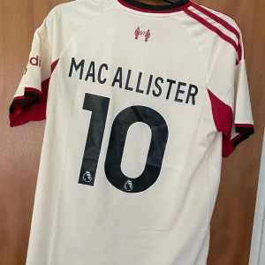 Liverpool bortatröja Mac Allister L - Liverpool FC bortatröja från Adidas med Mac Allister och nummer 10 på ryggen. Tröjan är vit med röda detaljer, klassiska Adidas-ränder på axlarna och klubbmärke på bröstet. Tillverkad i lätt och ventilerande polyester med AEROREADY-teknologi.