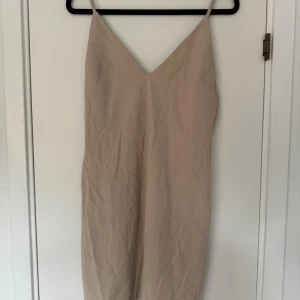 Beige klänning med smala axelband och djup rygg  - Stilren beige klänning med smala axelband och v-ringning framtill. Klänningen har en enkel, rak passform och dragkedja i ryggen samt djup i ryggen. Perfekt för dig som gillar minimalistisk stil och neutrala färger. Materialet känns lätt och svalt.