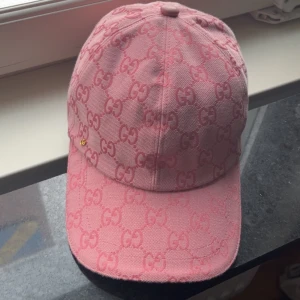 Rosa Gucci keps med GG-mönster - Säljer en rosa keps från Gucci med ikoniskt GG-mönster över hela kepsen. Justerbar rem baktill i matchande rosa och klassisk böjd skärm. Kepsen är tillverkad i bomull och har en exklusiv känsla, perfekt för dig som vill sticka ut med en lyxig accessoar.