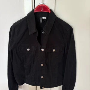 Svart jeansjacka från Gina Tricot - Svart jeansjacka från Gina Tricot med klassisk krage, silverfärgade knappar och två bröstfickor med lock. Jackan har lång ärm och en rak, något croppad passform. Perfekt för dig som vill ha en stilren och enkel look.