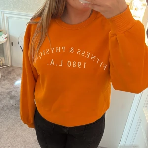 Orange sweatshirt från Gina Tricot - Cool orange sweatshirt från Gina Tricot med vit text 'Fitness & Physique 1980 L.A.' framtill. Tröjan har rund hals, långa ärmar och en relaxed passform. Perfekt för dig som gillar färg och vill sticka ut lite extra. Mjuk insida och ribbade muddar. 