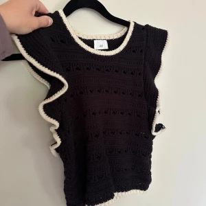 Svart virkad topp med volang från H&M - Svart virkad topp från H&M med kontrasterande vita kanter. Toppen är ärmlös med snygga volangdetaljer längs axlarna och nederkanten. Perfekt för dig som gillar en bohemisk och trendig stil. Materialet känns luftigt och mönstret är genombrutet för en cool look.