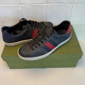 Gucci Sneakers - Snygga svarta Gucci sneakers med klassiskt GG-mönster och röd/blå rand på sidan. Skorna har svarta skosnören, vit sula och detaljer i rött och blått på hälen. Tillverkade i canvas med skinn-detaljer. Perfekta för dig som vill sticka ut med lyxig streetstyle. Perfekt skick och pris kan förhandlas vid snabbaffär