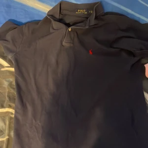 Mörkblå pikétröja från Polo Ralph Lauren - Klassisk mörkblå pikétröja från Polo Ralph Lauren i storlek S. Tröjan har korta ärmar, krage och två knappar framtill. Den ikoniska röda broderade loggan syns på bröstet. Tillverkad i bomull för en skön och stilren känsla.