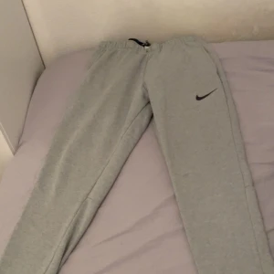 Grå mjukisbyxor från Nike - Snygga grå mjukisbyxor från Nike med svart logga på vänster ben. Byxorna har elastisk midja med snörning och är tillverkade i mjukt bomullsmaterial. Perfekta för chill eller träning, med en enkel och stilren look.