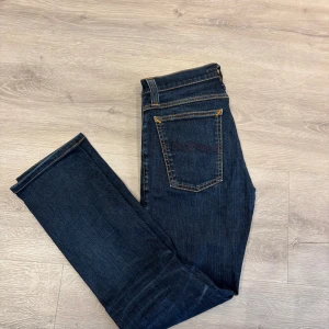 Mörkblå jeans från Nudie Jeans Co W26 L32 - Snygga mörkblå jeans från Nudie Jeans Co med klassisk femficksdesign, kontrastsömmar och läderpatch bak. Jeansen har rak passform och är tillverkade i slitstarkt denimtyg. Perfekta för dig som gillar stilrena och tidlösa jeans.