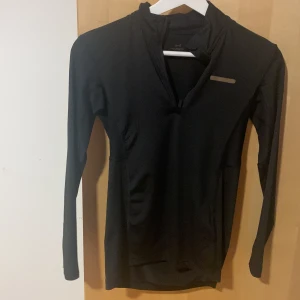 Svart långärmad träningströja A-Z - Svart långärmad träningströja från A-Z med halv dragkedja framtill och diskret reflexdetalj på bröstet. Tillverkad i ett stretchigt syntetmaterial som andas, perfekt för löpning eller gym. Snygg och funktionell passform med hög krage.