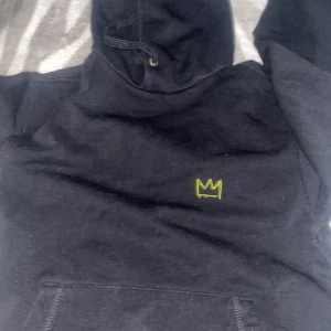 Svart hoodie med gul krona och text - Svart hoodie från Lucille med gul krona broderad på bröstet och unik mindmap-tryck i gult och lila på ryggen. Tröjan har huva med snörning och en stor magficka. Perfekt för dig som gillar streetwear och coola detaljer.