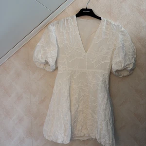 Vit kortklänning med puffärm - Supersöt vit kortklänning från KappAhl med broderade blommor och puffiga korta ärmar. Klänningen har v-ringad hals och voluminös kjol. Materialet känns lätt och har en snygg struktur tack vare broderierna. Den har används en gång. Det är oklart vart den är ifrån då jag köpte den begagnad men den var oanvänd. Den har en liten fläck längst ner men går kanske bort med starkare medel. (Sista bilden) 