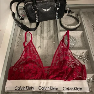 Spets bh Calvin Klein - Vinröd bh från Calvin Klein i spets med blommigt mönster. Elastiskt vitt band med logga runt om, smala axelband och triangelformade kupor. Storlek 70B.