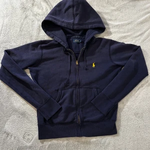 Marinblå hoodie från Polo Ralph Lauren - Säljer en marinblå hoodie från Polo Ralph Lauren med dragkedja och huva. Tröjan har klassisk gul logga broderad på bröstet, känguruficka och ribbade muddar.  Skick se bilder