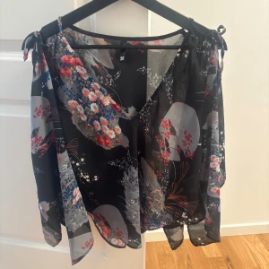 Blommig omlottblus i svart och grått - Säljer en luftig och transparent omlottblus med blommigt mönster i svart, grått, rött, blått och vitt. Blusen har knytband på axlarna och vida ärmar för en flowy känsla. Perfekt för dig som gillar unika detaljer och färgglada prints.