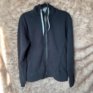 Blå hoodie från James Harvest, L - Blå hoodie från James Harvest i storlek L. Tröjan har dragkedja framtill, två fickor med dragkedja, huva med ljusa snören