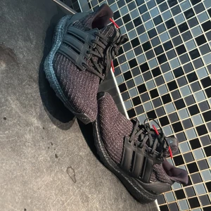 Adidas Ultraboost svarta sneakers - Svarta Adidas Ultraboost sneakers med stickad ovandel och svarta plastdetaljer på sidorna. Skorna har svarta snören med röda spetsar och röd innersula med BOOST-tryck. Snygg, chunky sula och klassisk tre-randig design på sidan.