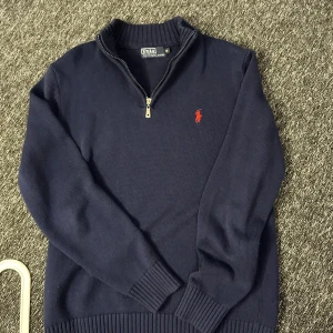 Mörkblå stickad tröja Polo Ralph Lauren - Mörkblå stickad tröja från Polo Ralph Lauren med halv dragkedja och röd broderad logga på bröstet. Tröjan har ribbade muddar och hög krage, perfekt för kyliga dagar. Klassisk och stilren modell som funkar till det mesta.