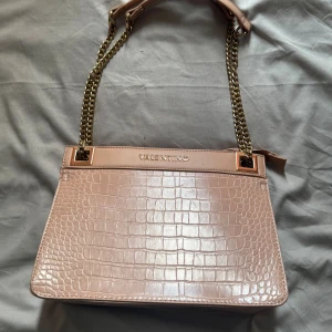 Rosa väska från Valentino - Snygg rosa väska från Valentino med krokopräglat mönster och guldfärgade kedjor. Väskan har dragkedja upptill och detaljer i guld. Perfekt storlek.