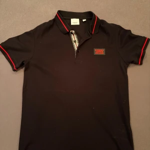 Svart Burberry pikétröja med röd detalj - Svart pikétröja från Burberry med röd logotyp på bröstet och röda ränder på krage och ärmslut. Klassisk krage med knappar och snyggt mönstrad knappslå. Perfekt för dig som vill ha en stilren och exklusiv look. Om du är intresserad TVEKA INTE på att höra av dig… helt oanvänd 