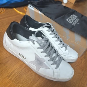 Golden Goose vita sneakers med stjärna - Golden Goose sneakers i vitt skinn med grå mockadetaljer och ikonisk stjärna på sidan. Skorna har grå snörning, svart hälkappa och platt sula. Perfekta för dig som gillar en clean men ändå unik look. Kommer med originalkartong och dustbag.