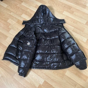 Svart Moncler pufferjacka med huva - Säljer en svart, glansig pufferjacka från Moncler med huva och klassisk logga på ärmen. Jackan har dragkedja, flera fickor och quiltad design. Perfekt för kalla dagar och riktigt snygg streetstil.