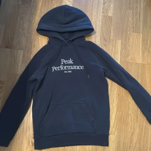 Mörkblå hoodie från Peak Performance - Säljer en mörkblå hoodie från Peak Performance med vit broderad logga på bröstet. Tröjan har huva med snörning, känguruficka och ribbade muddar. Perfekt för chill dagar eller när du vill ha en sportig look.