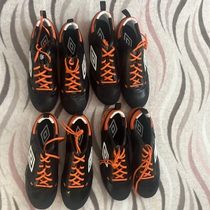 Umbro svarta och orangea fotbollsskor - Säljer 4 par Umbro fotbollsskor i svart och orange med vita detaljer. Skorna har snörning, rund tå och platt sula med dobbar för bra grepp på konstgräs. Snygg sportig design med märkets logga på sidan och kontrasterande orange snören. Säljer skorna eftersom jag inte längre behöver dem. Kunden får alla par för 800 kr. De finns i storlek ett par stl 43, 44 och 2 två par i 44,5. De är även i nytt skick har aldrig användt dem.