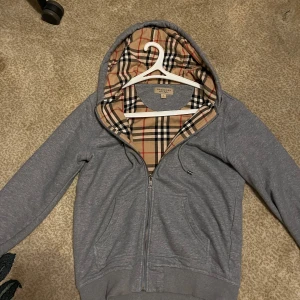 Grå hoodie från Burberry med rutig huva - Säljer en grå hoodie från Burberry med dragkedja och klassiskt rutigt foder i huvan. Tröjan har långa ärmar, snörning i huvan och ribbade muddar. Perfekt för dig som vill ha en stilren men ändå lyxig look.