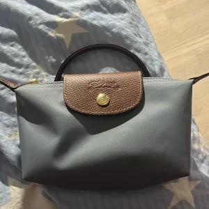 Grå handväska från Longchamp - Snygg grå handväska från Longchamp med bruna skinnhandtag och lock. Väskan har guldfärgade metalldetaljer och dragkedja med Longchamp-logga. Perfekt storlek för det viktigaste och klassisk design som aldrig går ur tiden.